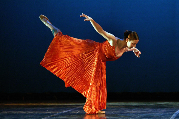 Natalie Lissack performing in an orange pleated dress